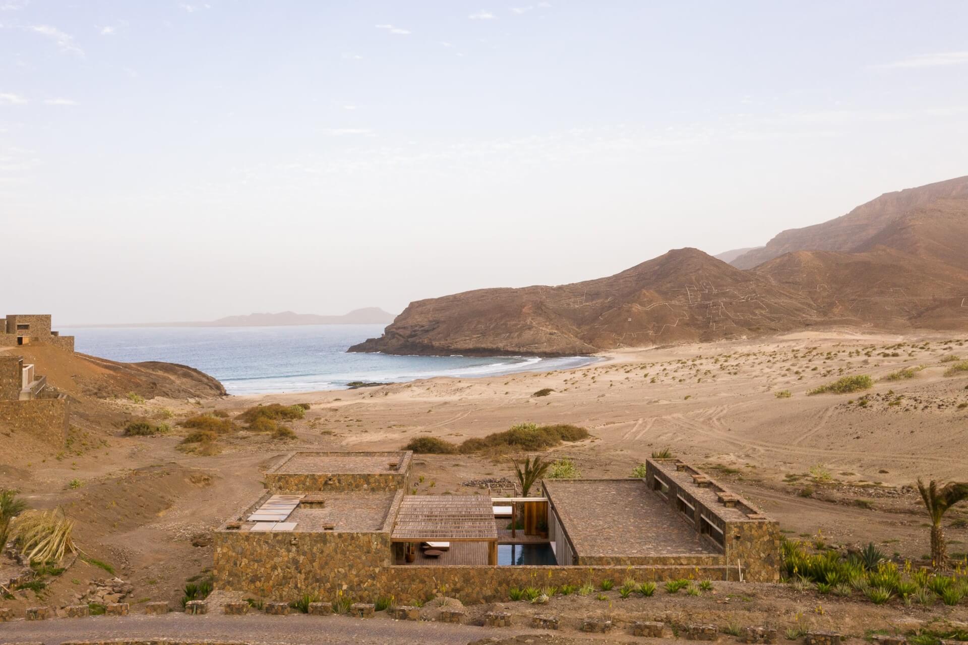 Barefoot Luxury, Africa — Minimalist stone pavilion on empty beach with volcanic headland, turquoise water, and desert landscape at dusk.