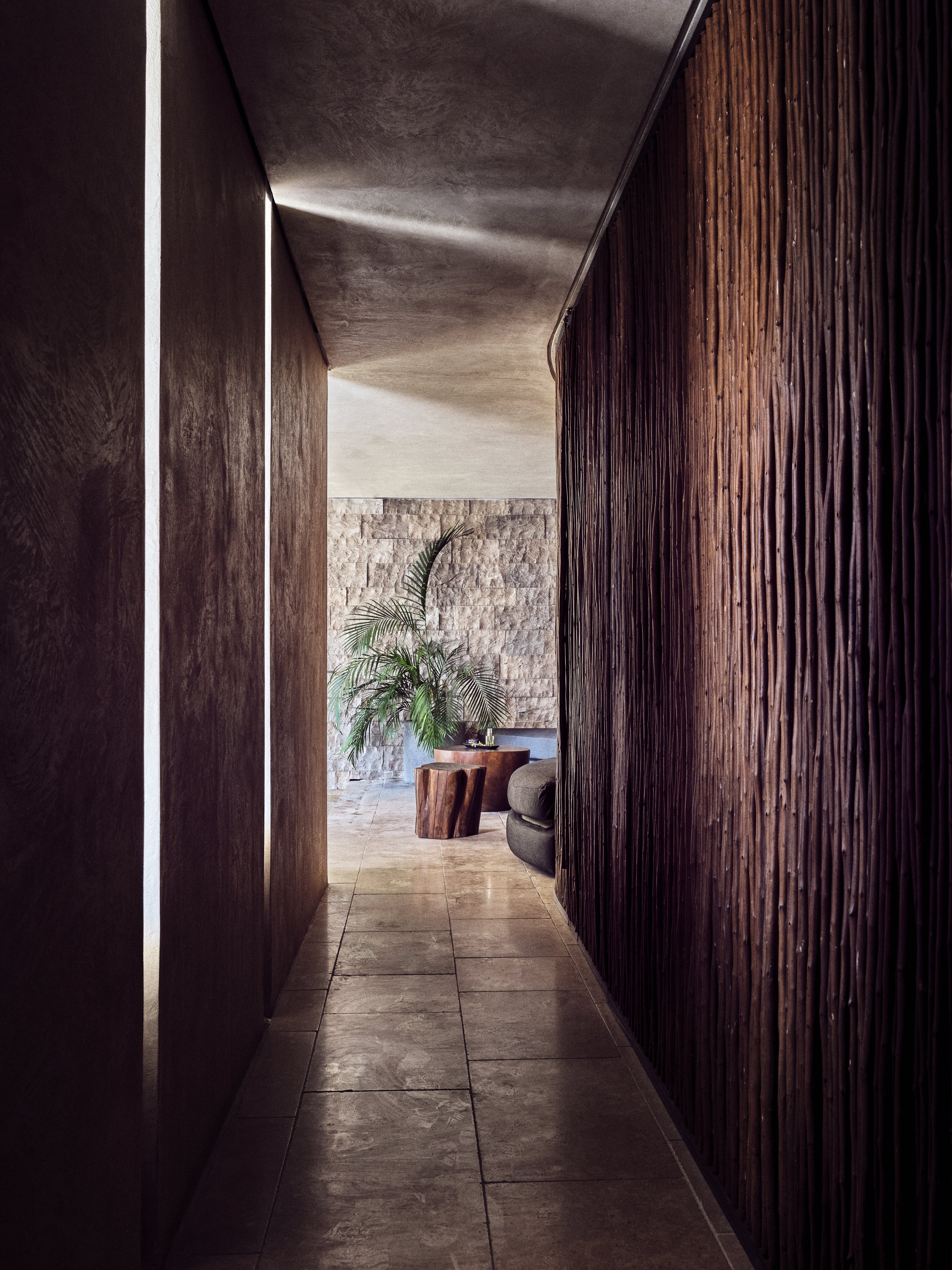 Olea All Suite Hotel, Europe — Dark timber corridor with angled ceiling, stone flooring, opening to courtyard with potted palms and textured stone wall.