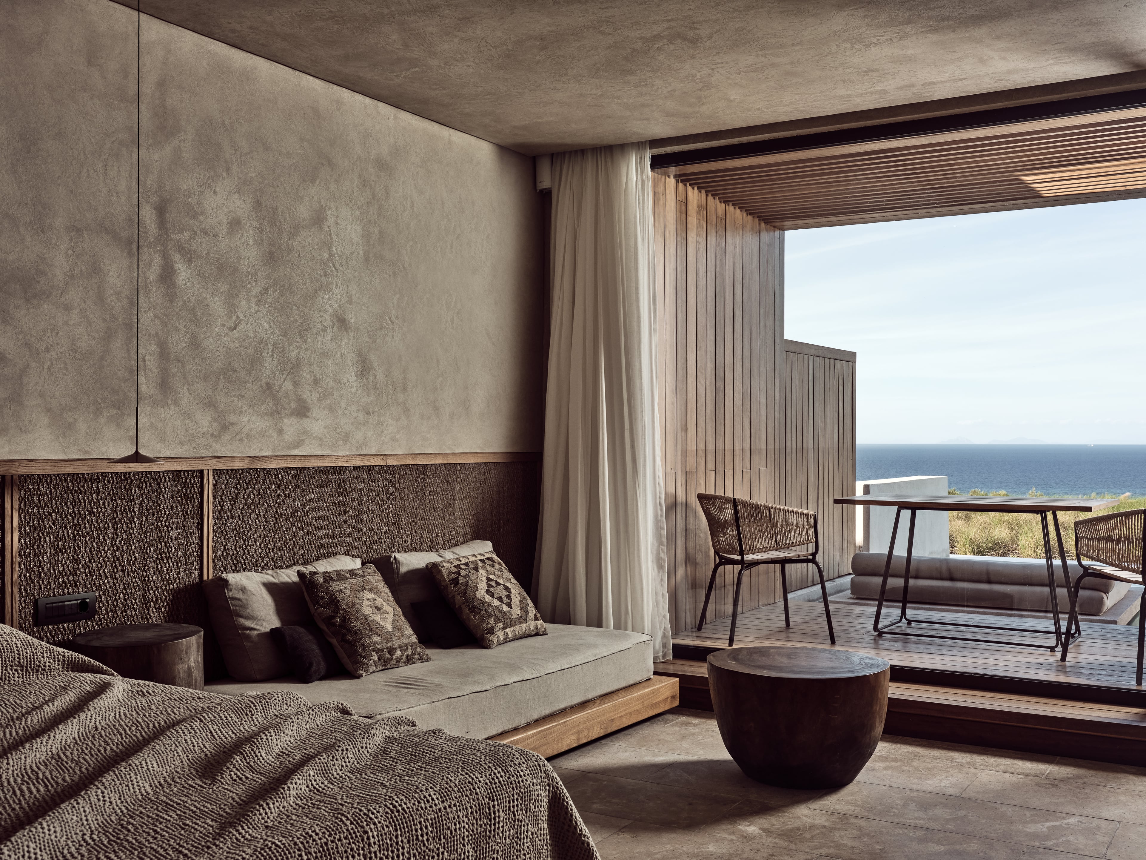Olea All Suite Hotel, Europe — Coastal bedroom with woven headboard, linen seating, concrete ceiling, and panoramic sea views from private terrace.