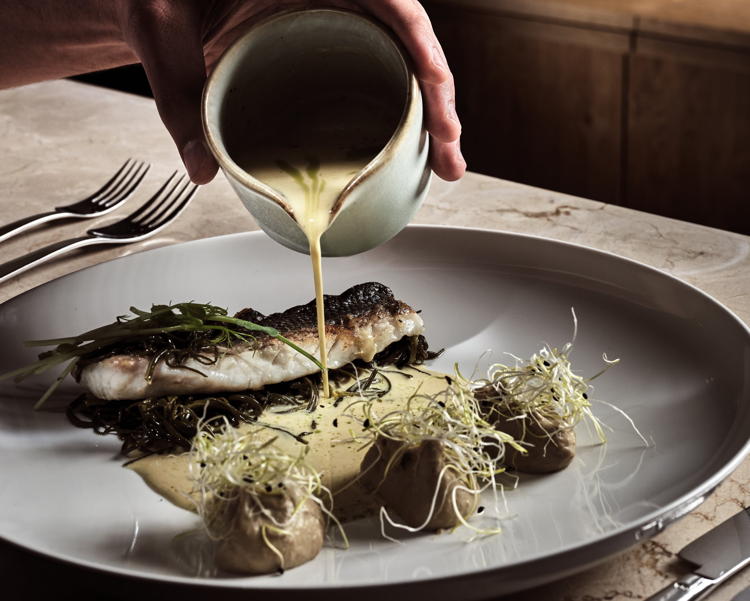 Olea All Suite Hotel, Europe — Chef pouring beurre blanc sauce over seared fish fillet with seaweed and microgreens on white plate, fine dining presentation.
