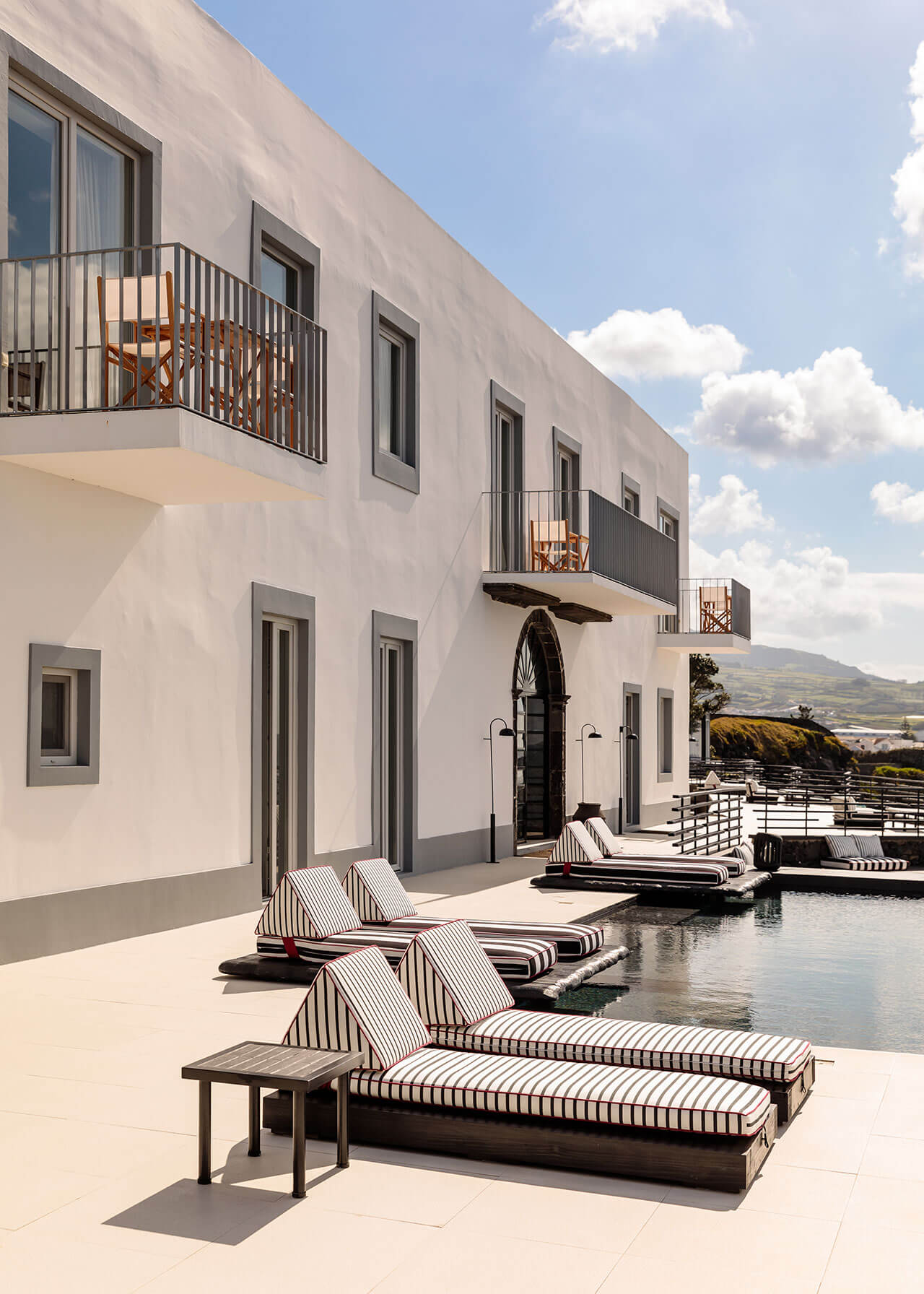 White Hotel, Europe — Modernist waterfront villa with striped loungers on limestone terrace, minimalist white facade, black balcony railings, mountain views under clear sky.