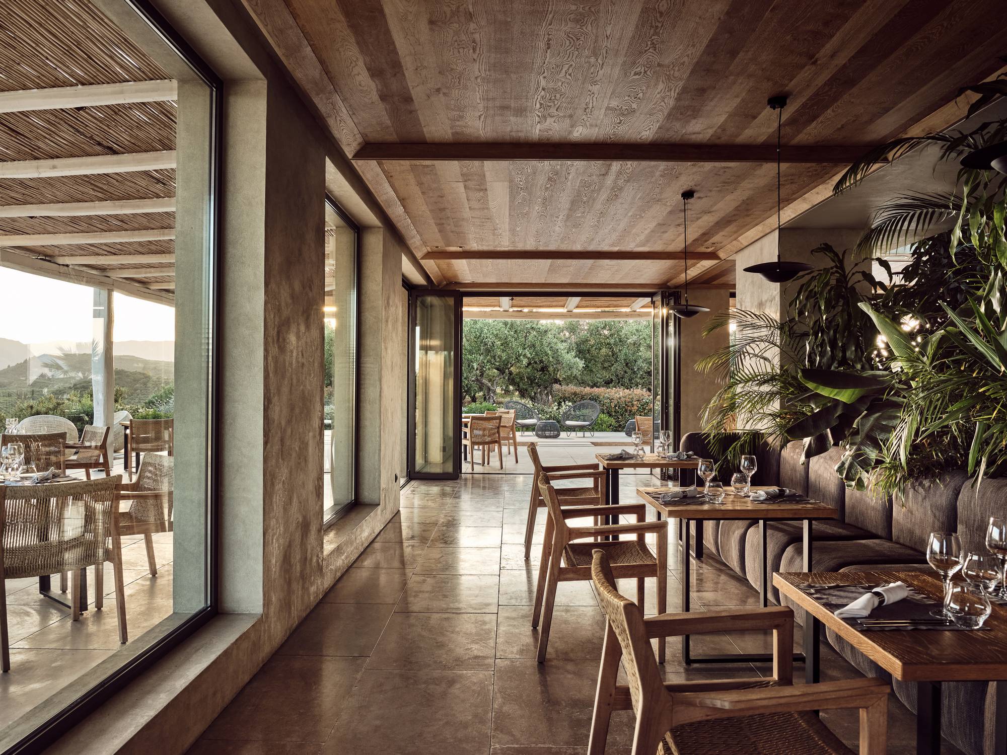Flow Dine & Wine, Europe — Modern dining room with timber ceiling beams, concrete walls, natural light through floor-to-ceiling windows, wooden chairs, potted plants, and terrace views.