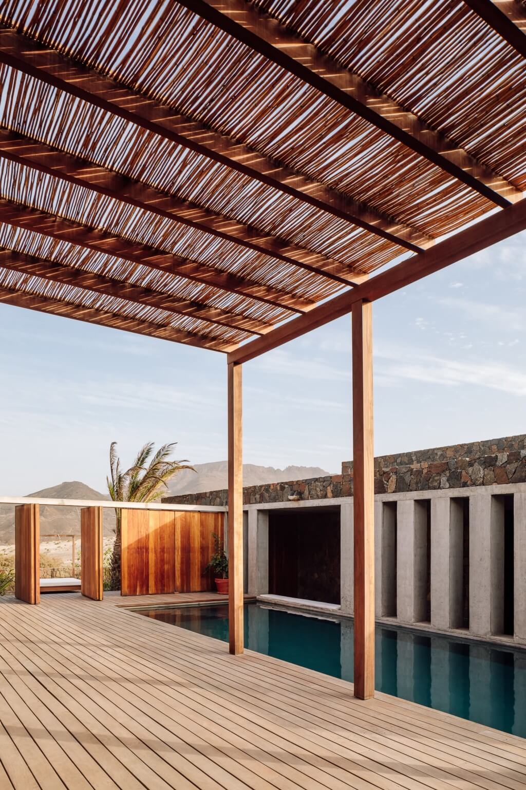 Barefoot Luxury, Africa — Wooden pergola with slatted roof casting linear shadows over teak deck and infinity pool, minimalist architecture with volcanic stone and concrete details overlooking desert landscape.