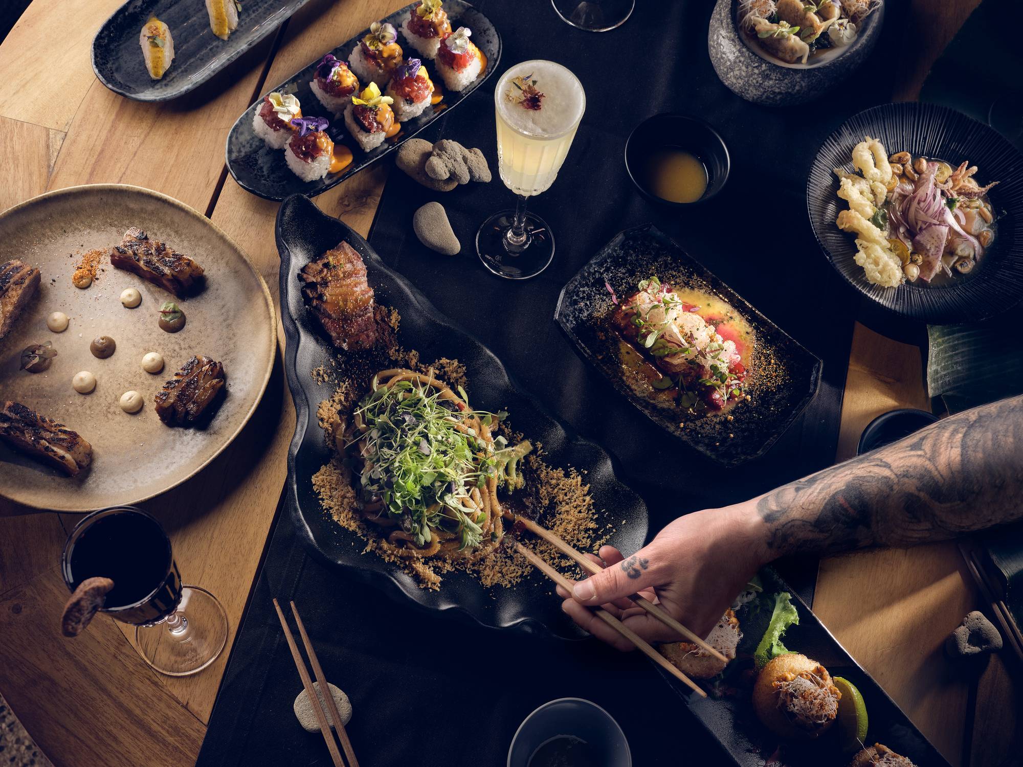 Pacifico by Nikkie, Europe — Overhead view of plated Asian cuisine with artistic presentation, dark tables, chopsticks, and tattooed hand reaching across modern fine dining setup.