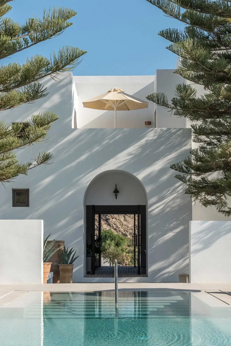 Parīlio Hotel, Europe — Minimalist white-washed courtyard with arched black doorway, reflecting pool, beige umbrella, and pine trees casting geometric shadows under clear sky.