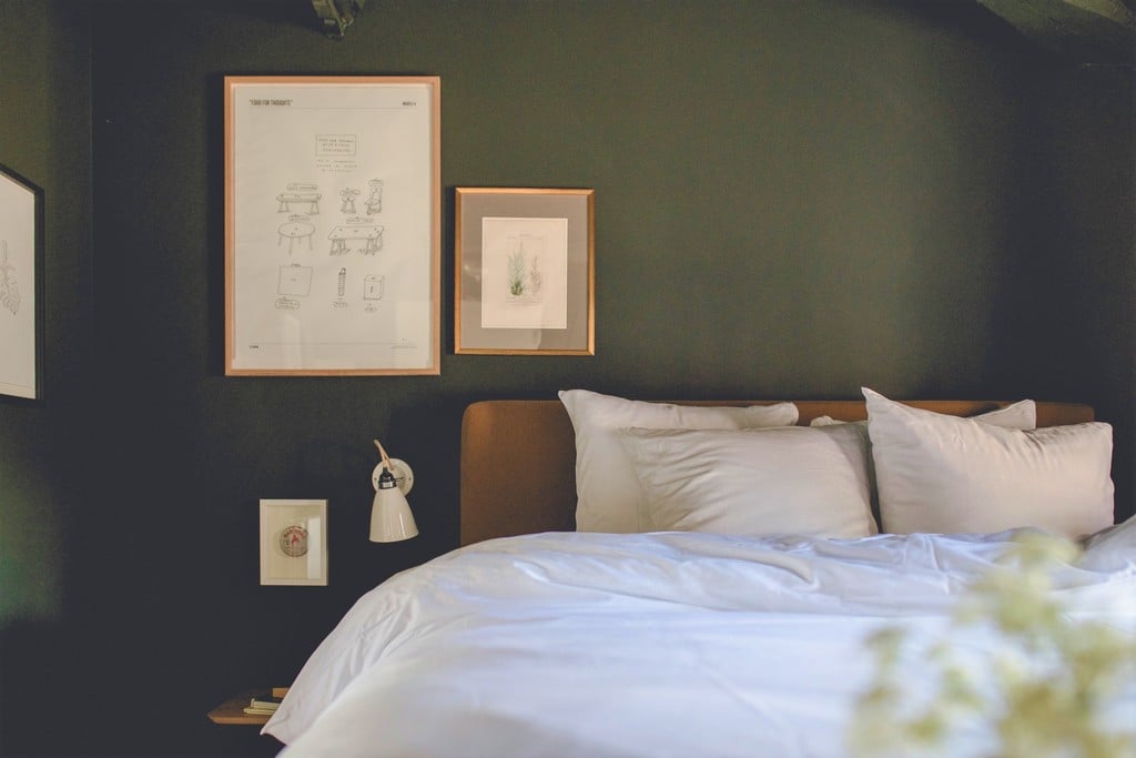 Le Barn, Europe — Dark green bedroom wall with wooden-framed botanical sketches, cognac leather headboard, white linens, and minimalist bedside styling.