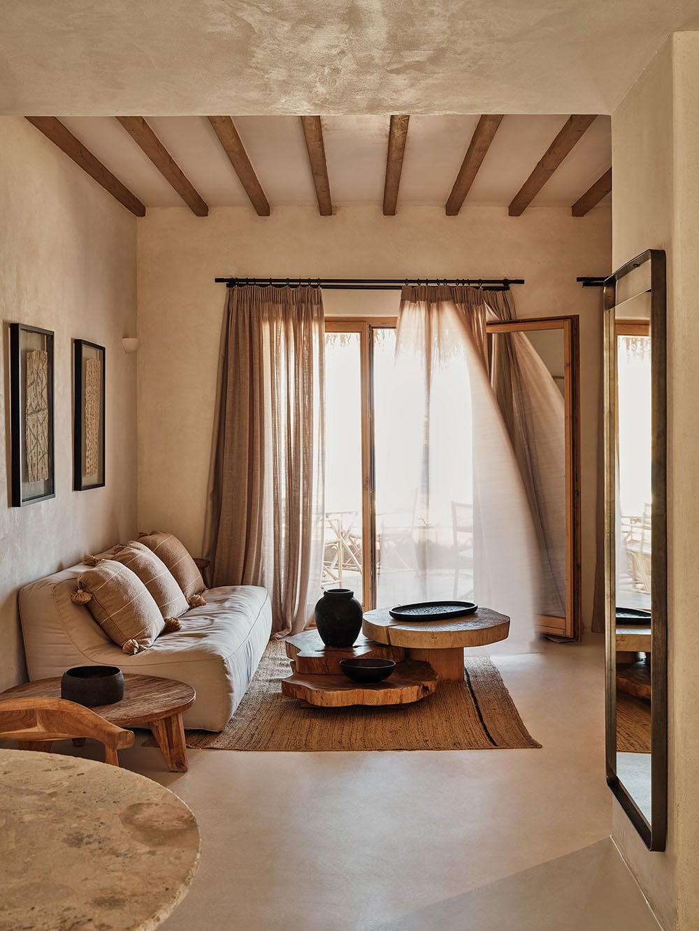 The Wild, Europe — Minimalist hotel bedroom with exposed wooden beams, cream plaster walls, linen daybed, wooden side tables, and sheer curtains filtering natural light through glass doors.