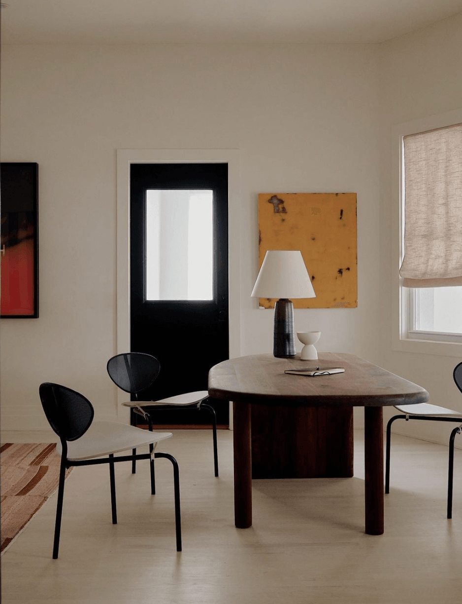 The James Bradley, North America — Minimalist dining room with oval dark wood table, black chairs, ceramic table lamp, and ochre artwork against cream walls.