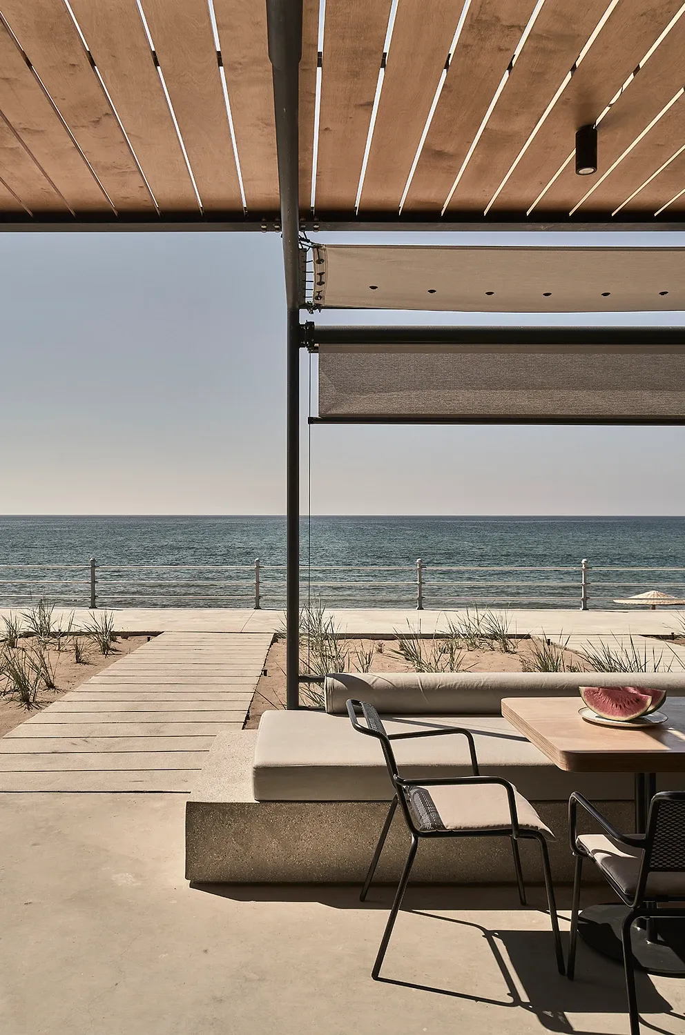 Dexamenes Seaside Hotel, Europe — Modern beachfront lounge with slatted wood pergola, concrete seating, ocean view, minimalist coastal architecture and natural light.