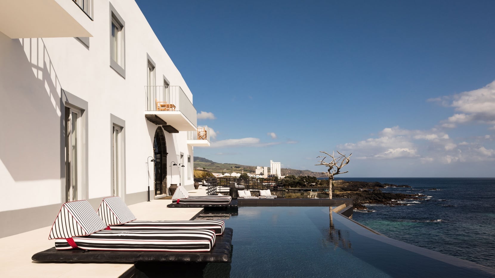 White Hotel, Europe — Striped loungers on infinity pool terrace overlooking coastal cliffs and ocean, modernist white architecture with floor-to-ceiling windows, clear blue sky.