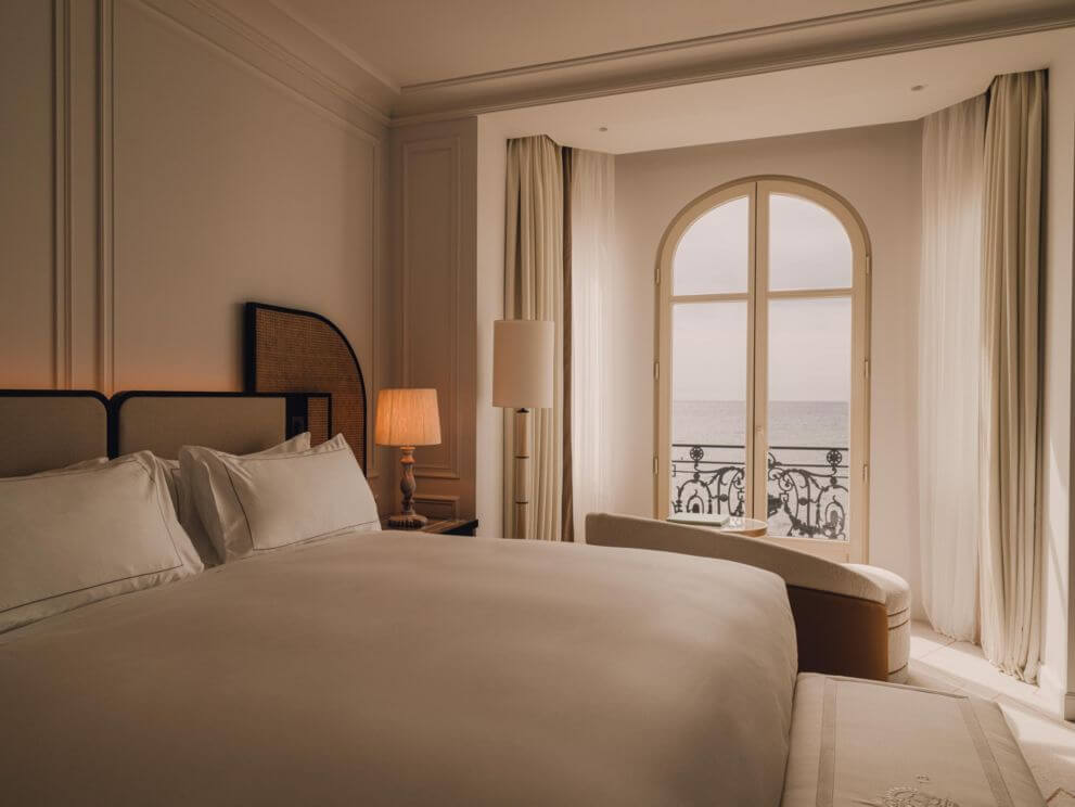 Carlton Cannes, Cannes, France — Minimalist bedroom with arched French doors, cream linens, rattan headboard, and Mediterranean sea view from wrought-iron balcony.
