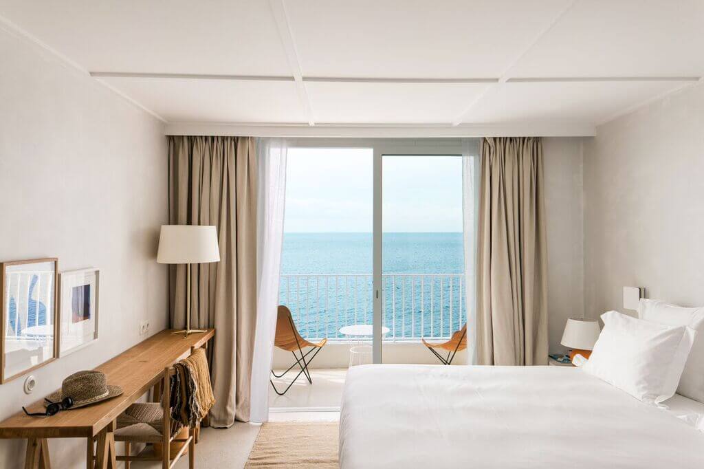 Les Roches Rouges, Europe — Minimalist beachfront bedroom with white bedding, wooden desk, tan linen curtains, and sea-view balcony doors.