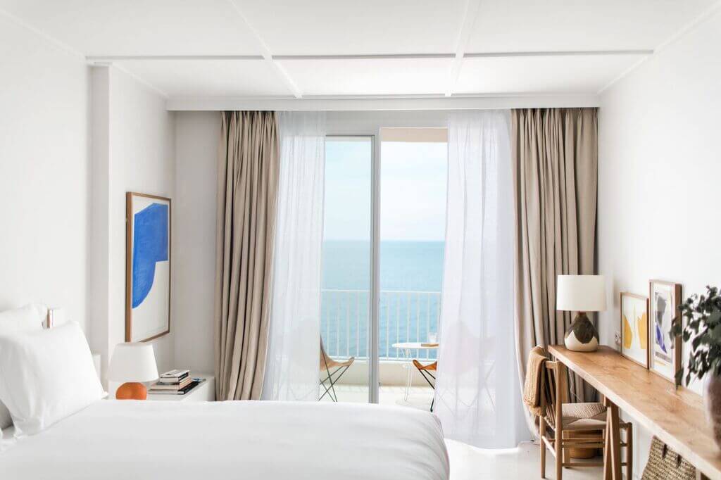 Les Roches Rouges, Europe — Minimalist coastal hotel bedroom with sea-view balcony, natural linen drapes, wooden desk, and modern neutral furnishings.