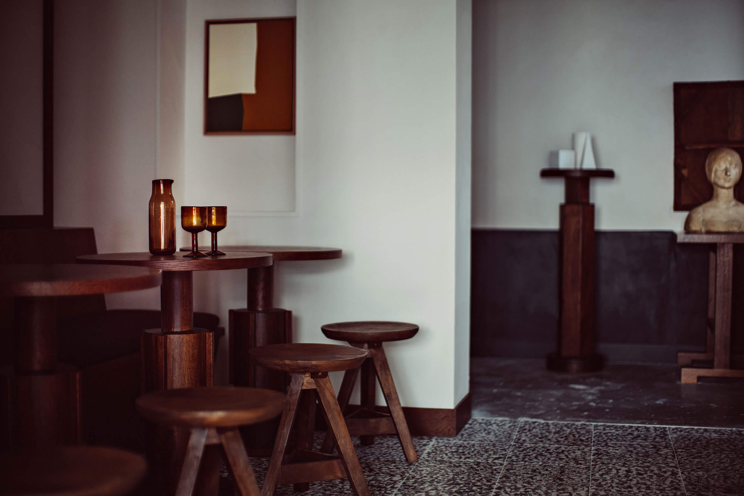 Académies et des Arts, Europe — Modernist lounge with dark wood furniture, amber glass vessels, geometric artwork, and sculptural bust on terrazzo floor.