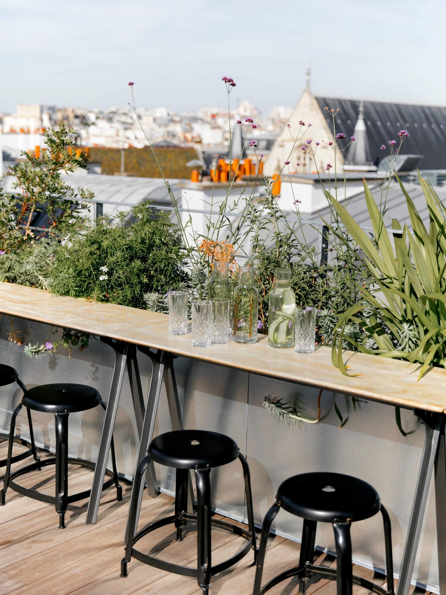 Hôtel National Des Arts, Europe — Rooftop bar with wood counter, black stools, potted flowers in glass vases, Parisian cityscape and church spire beyond.