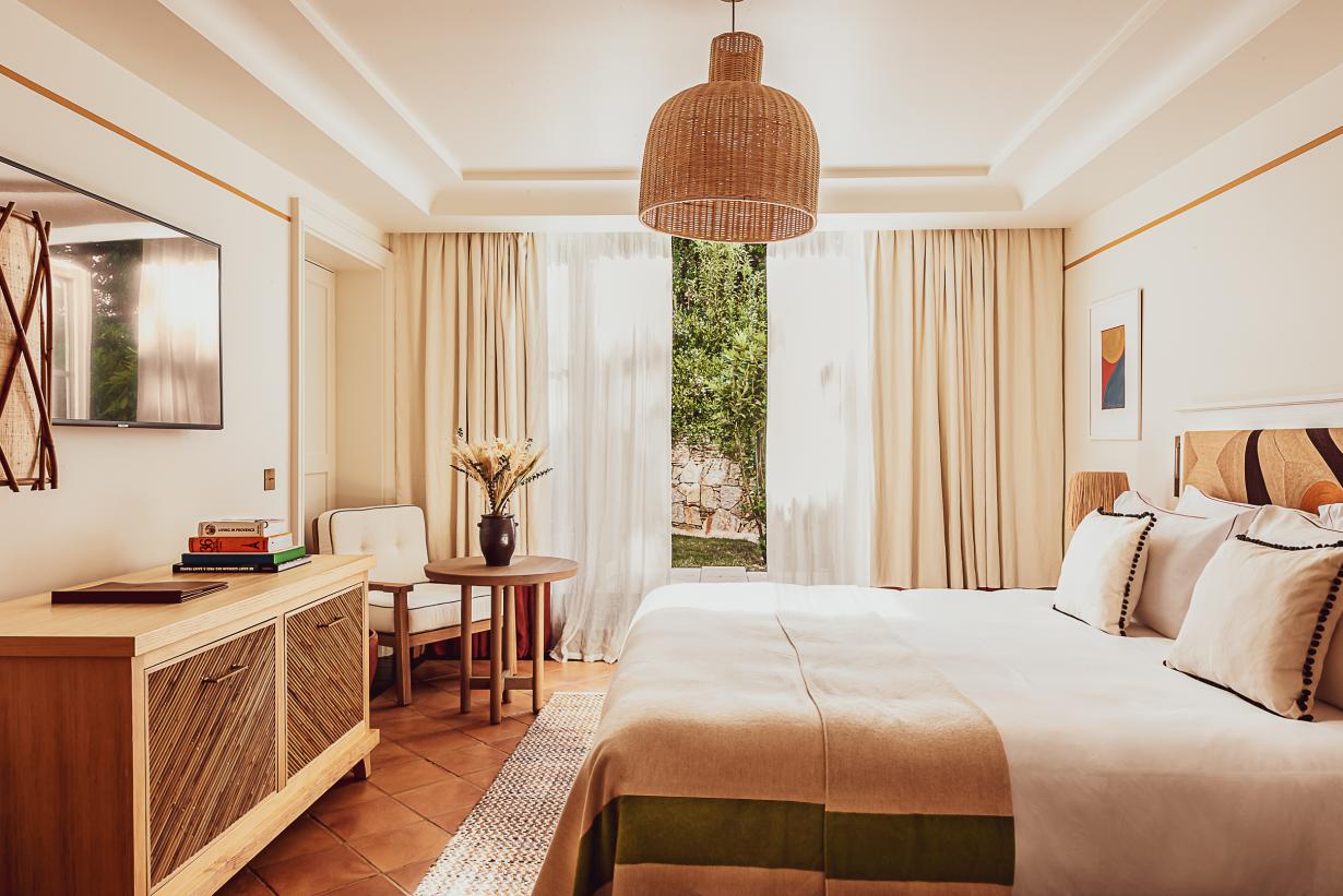 Hôtel Lou Pinet, Europe — Sunlit bedroom with woven pendant light, terracotta floors, cream linens, wooden furniture, sheer curtains, garden views, warm minimalist aesthetic.