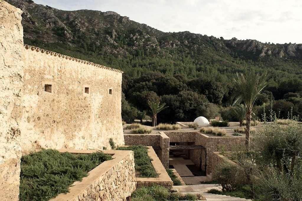 Es Racó d'Artà, Balearic Islands, Spain — Stone fortress with narrow windows overlooking terraced gardens, palm trees, and rocky Mediterranean hills in golden afternoon light.
