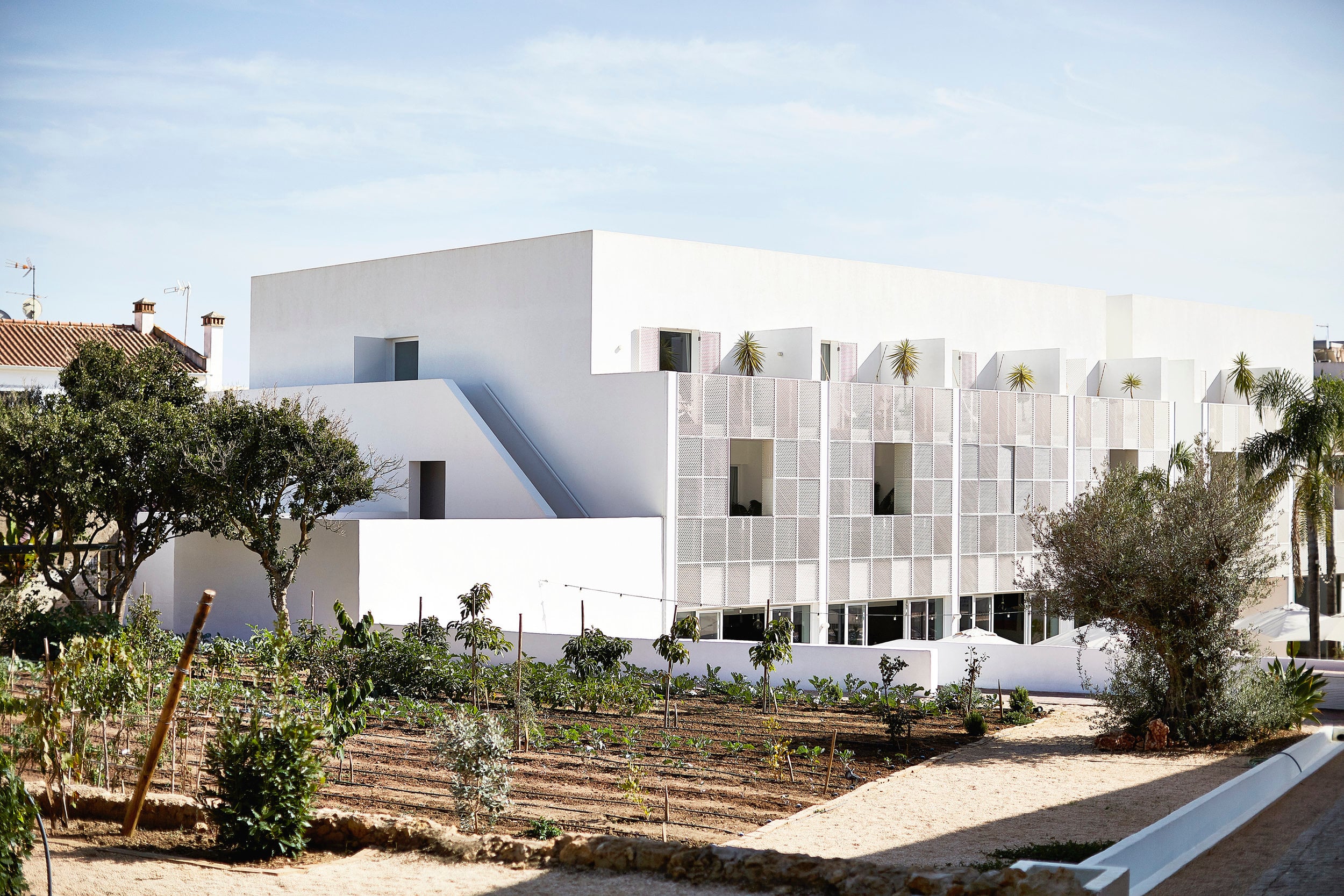 Casa Mãe, Europe — Modernist white building with perforated metal facade, geometric forms, and surrounding Mediterranean garden with olive trees under clear sky.