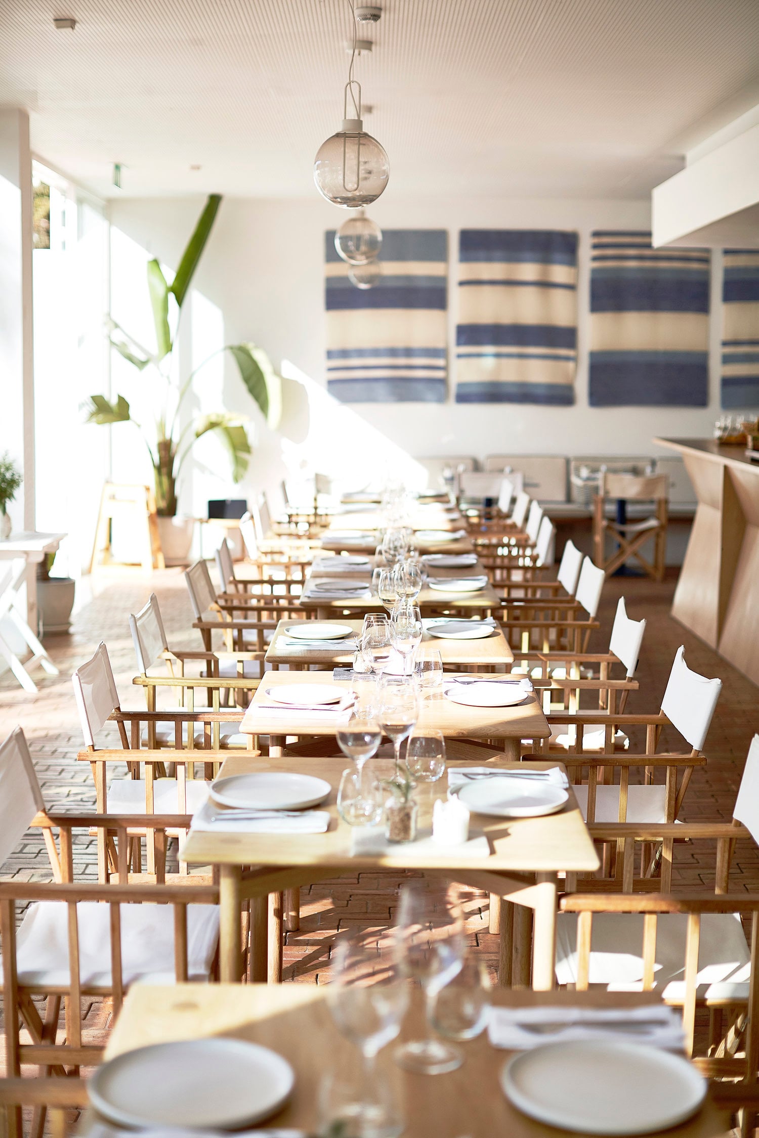 Orta Restaurant, Europe — Minimalist dining room with natural light, wooden chairs, striped textile wall art, globe pendant, and long communal table settings.