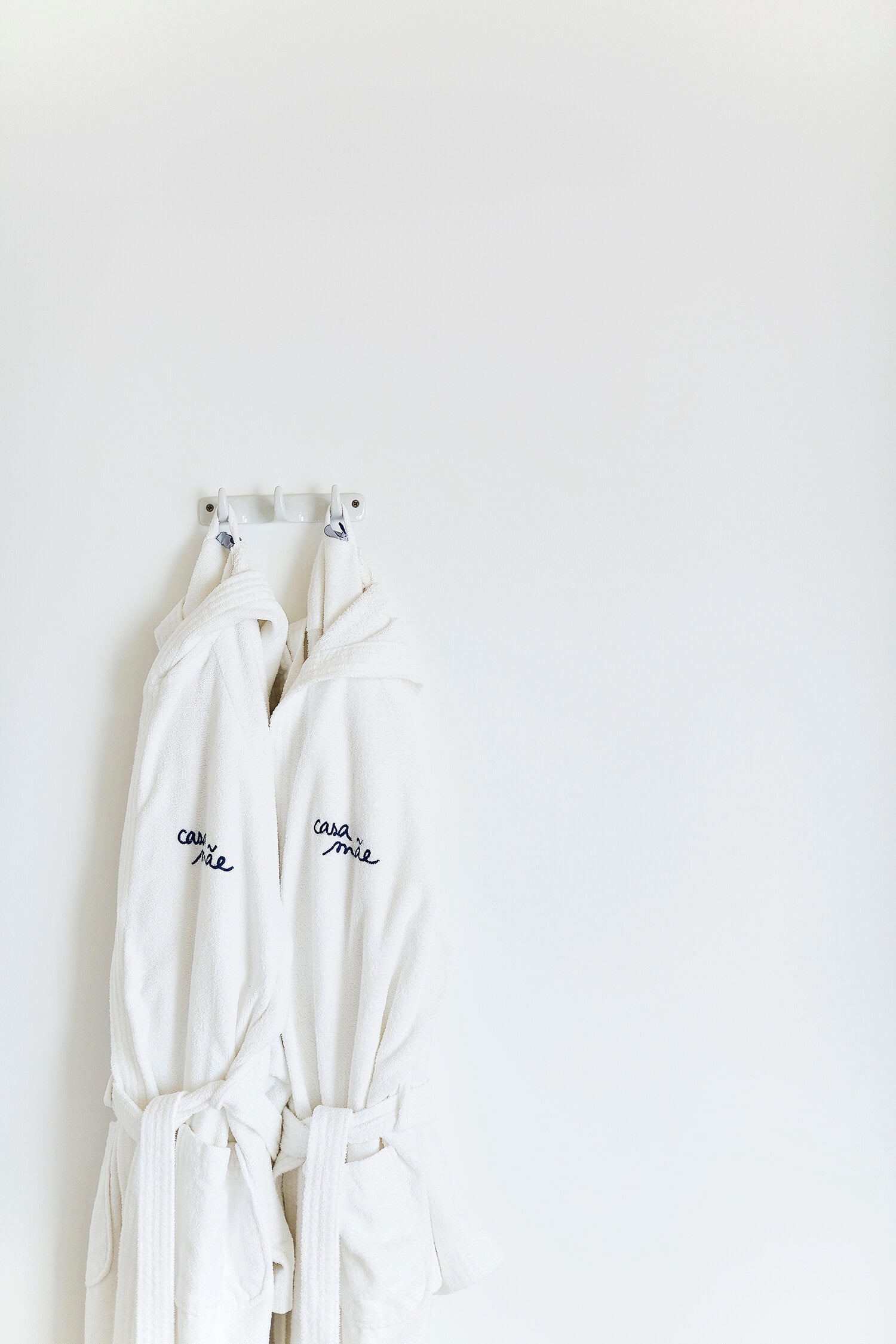 Casa Mãe, Europe — Two white linen robes with embroidered branding hang from wall hooks against minimalist interior, displaying luxury hospitality design detail.