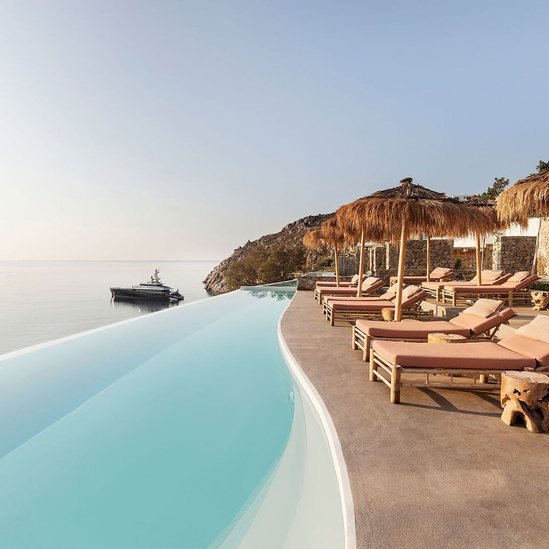 The Wild, Europe — Infinity pool with turquoise water overlooking Aegean Sea, wooden loungers beneath thatched umbrellas, stone architecture nestled into hillside, luxury resort aesthetic.