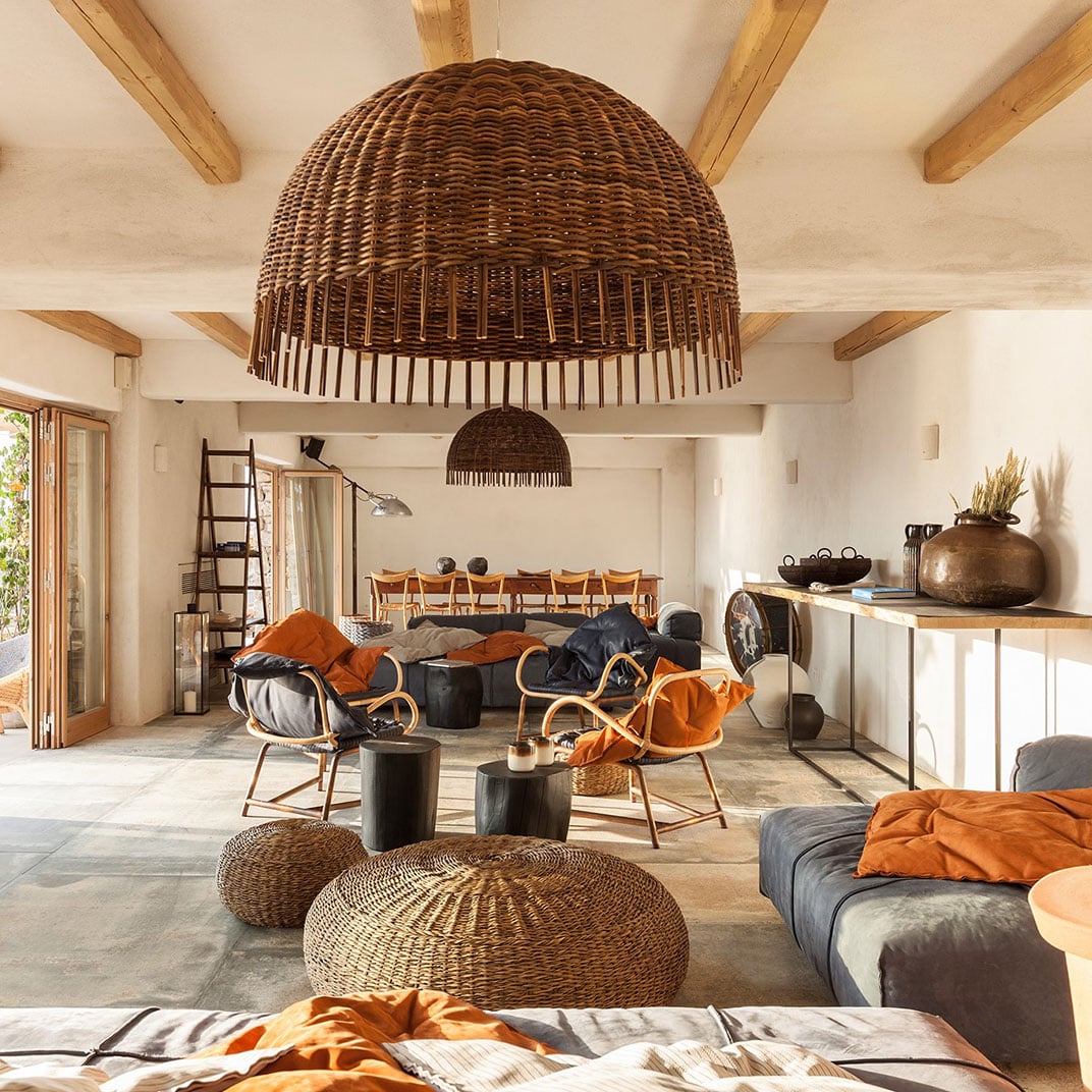 The Wild, Europe — Woven rattan dome pendant lights and wicker poufs in minimalist open-plan lounge with wooden beams, ochre accents, and natural stone flooring.