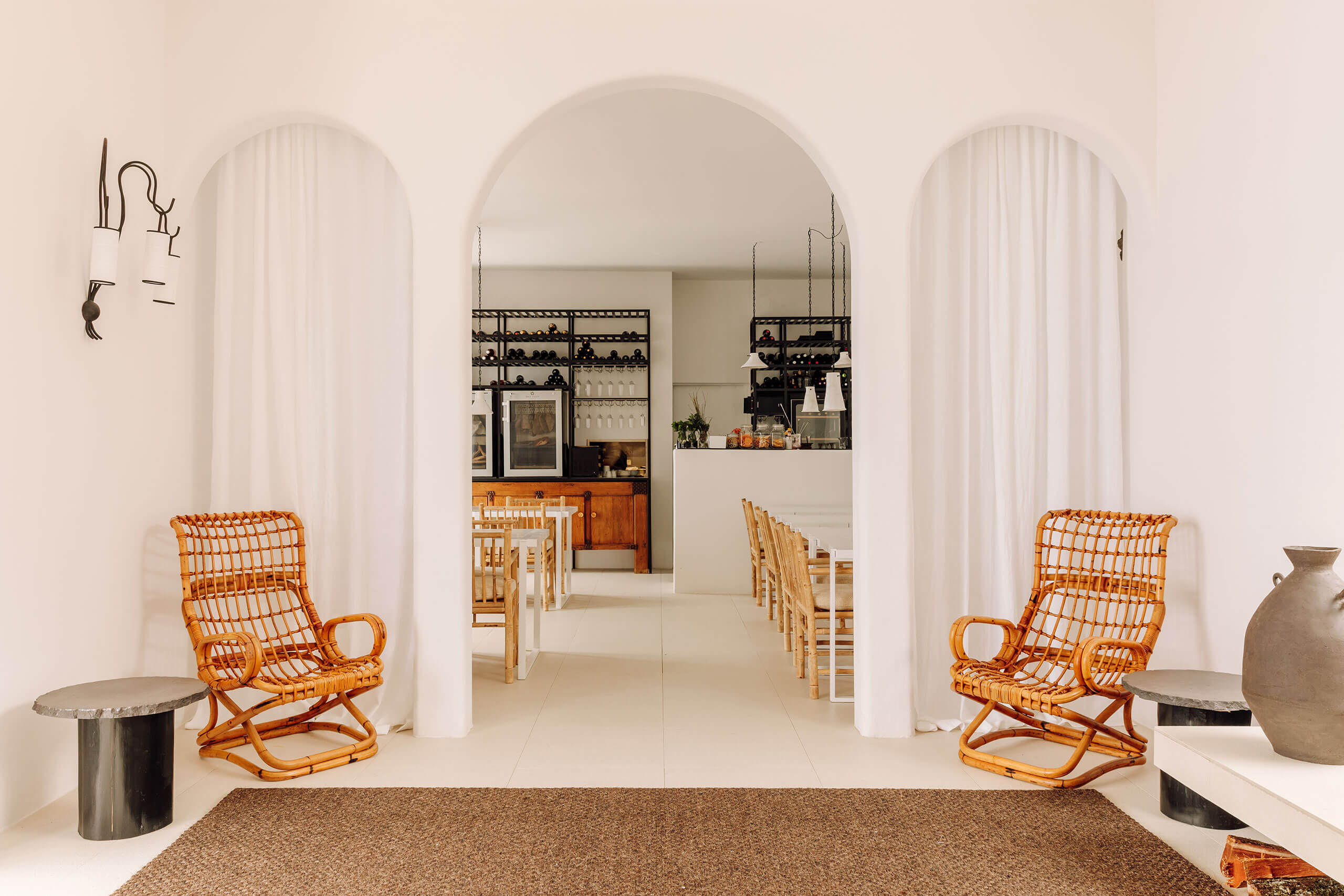 White Hotel, Europe — Minimalist lobby with arched doorways, rattan chairs, concrete side tables, and linen curtains opening to dining space.