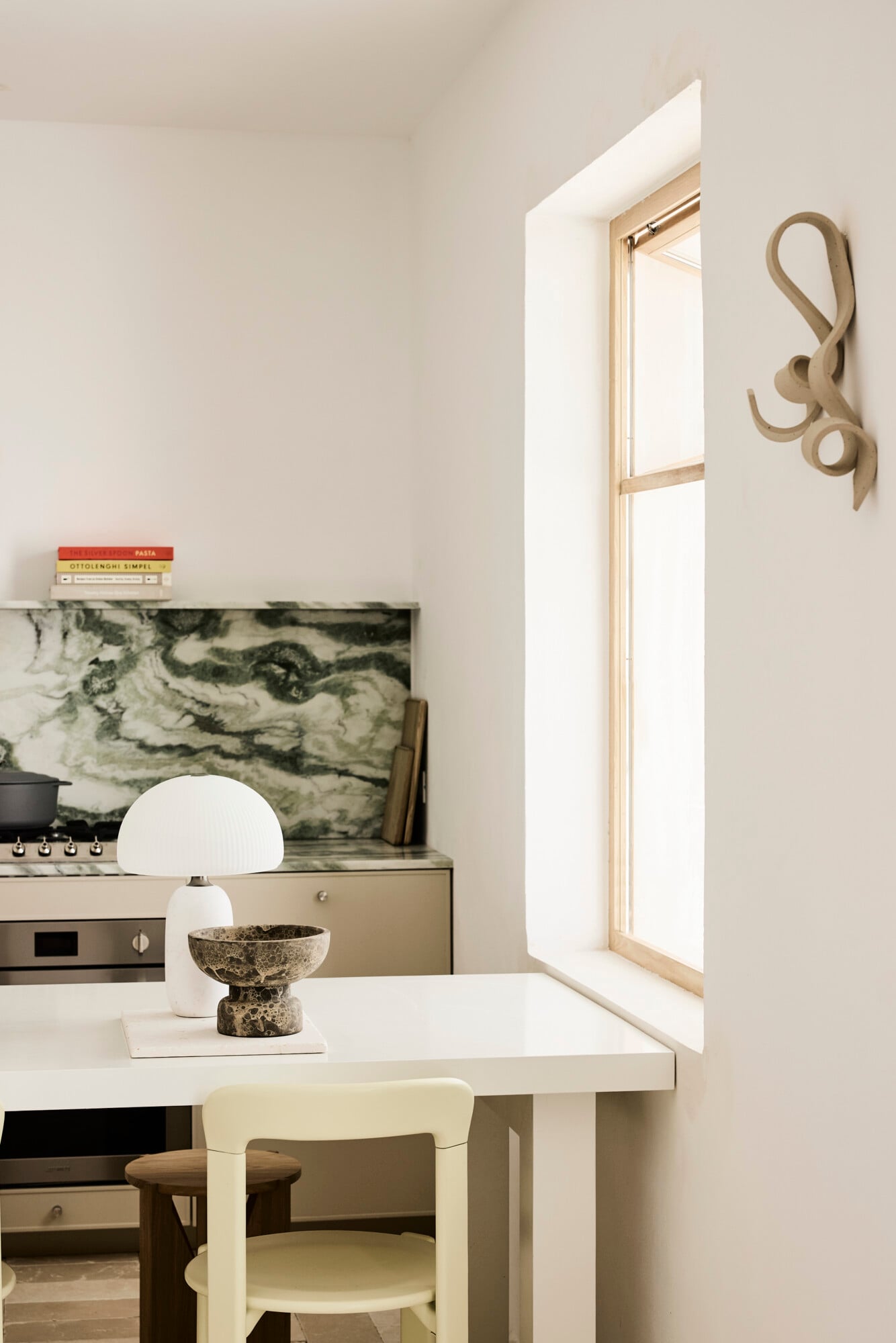 Villa Sessana, Italy — Minimalist kitchen with marble backsplash, natural light from tall window, white dome lamp, textured stone vessel on white counter.