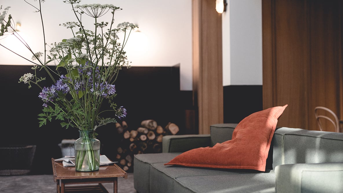 Le Barn, Europe — Modern hotel lounge with gray sofa, rust linen pillow, glass vase of wildflowers, wooden side table, and warm natural light through floor-to-ceiling windows.
