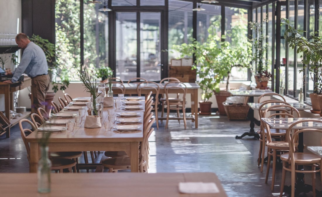 La Serre, Europe — Sunlit conservatory dining room with wooden tables, bentwood chairs, potted greenery, and floor-to-ceiling windows overlooking garden.