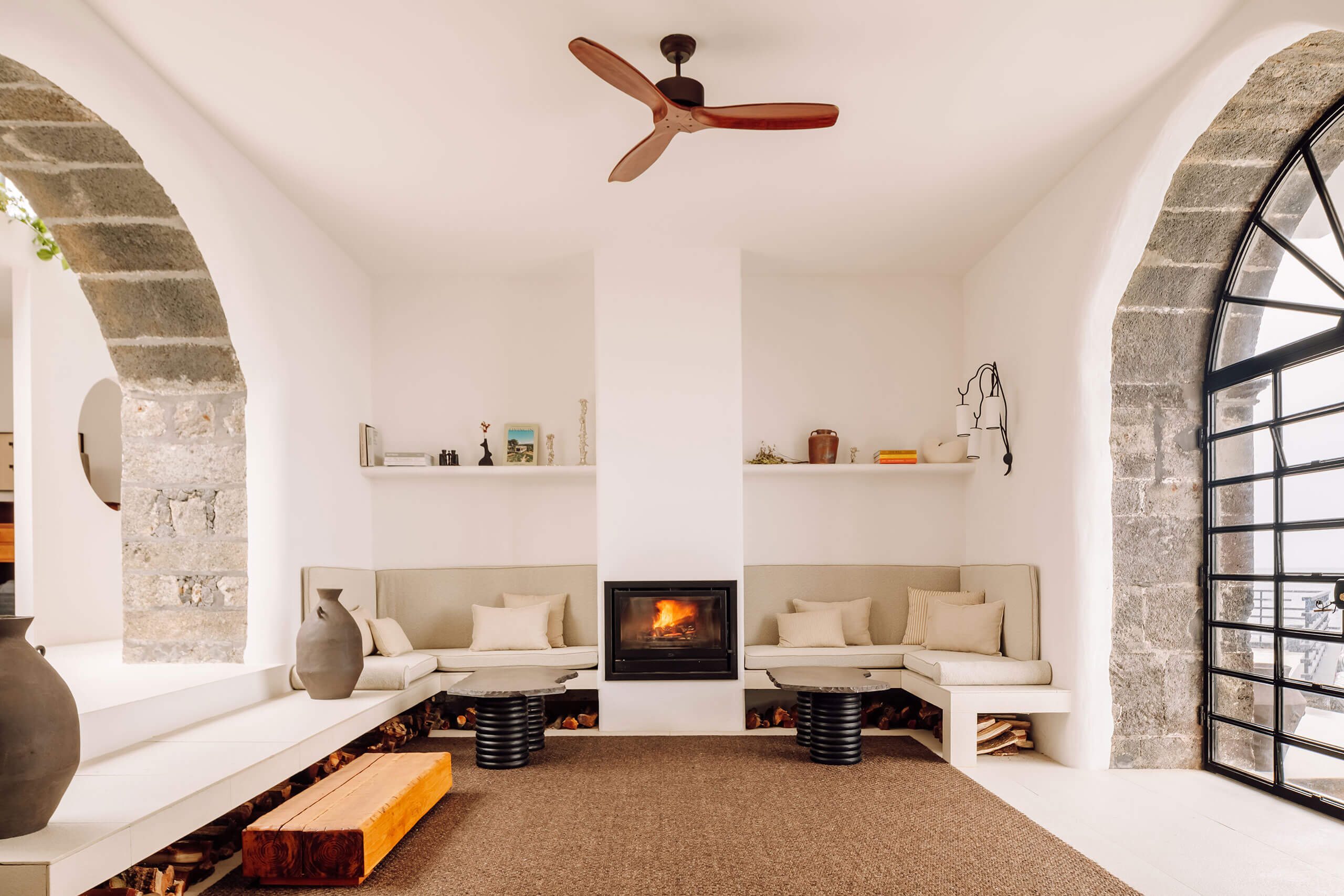 White Hotel, Europe — Minimalist lounge with curved stone archways, built-in seating, central fireplace, wooden ceiling fan, and natural light from grid-pattern windows.