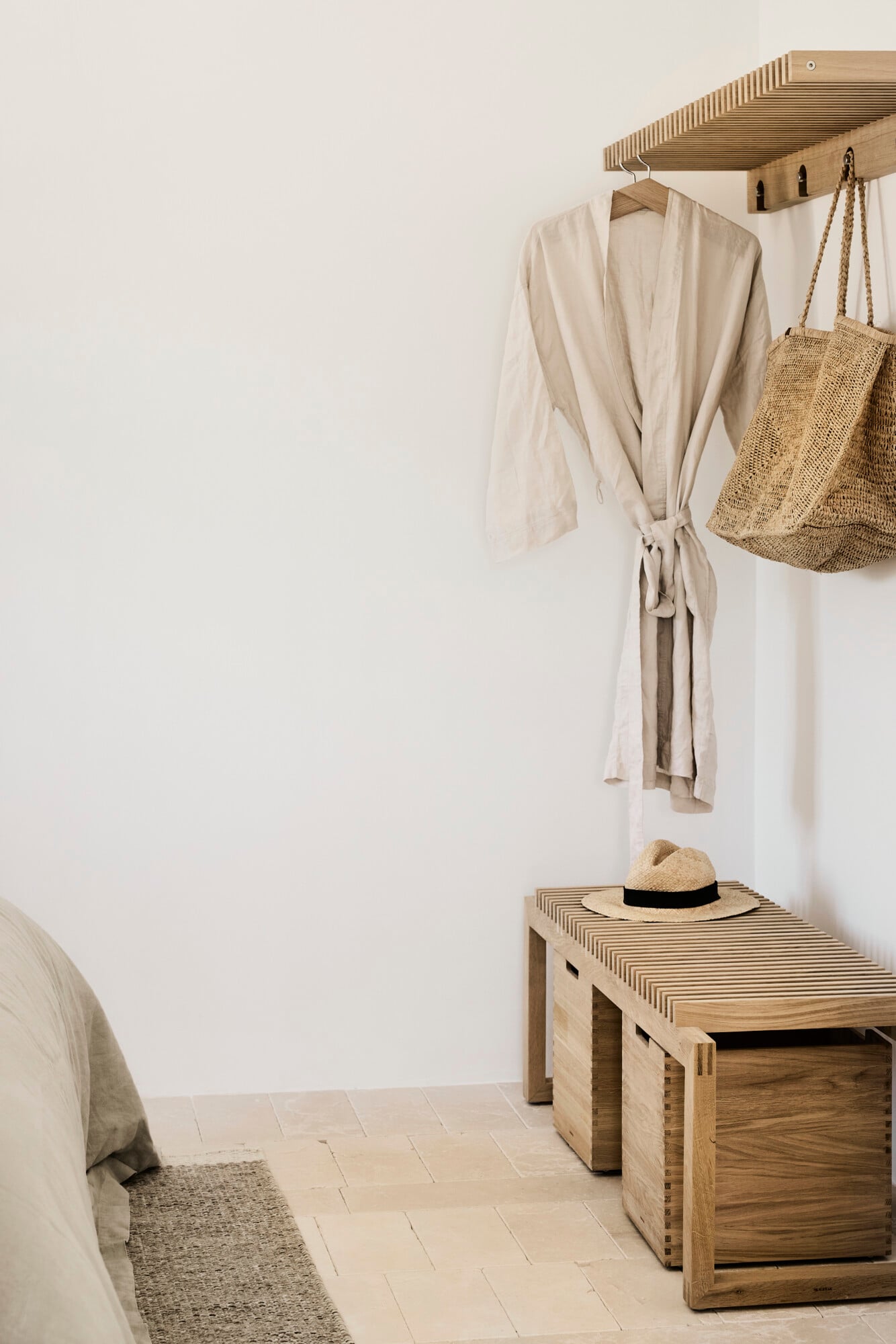 Villa Sessana, Italy — Minimalist bedroom corner with linen robe, woven tote bag, straw hat, wooden bench and storage, neutral palette, natural light.