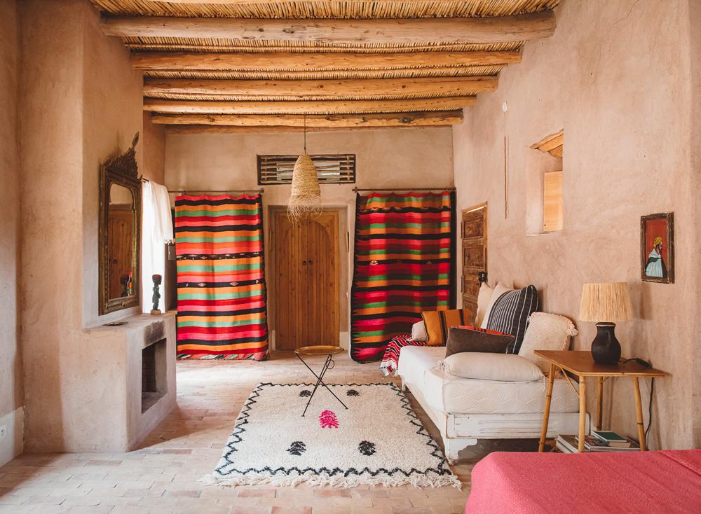Berber Lodge, Africa — Earthen-walled bedroom with exposed timber beams, vibrant striped textiles, woven pendant light, and traditional Berber furnishings.