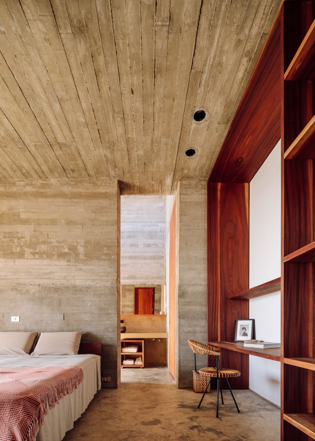 Barefoot Luxury, Africa — Concrete bedroom with timber ceiling, red wooden shelving, and woven chair at desk beside bed with coral throw.