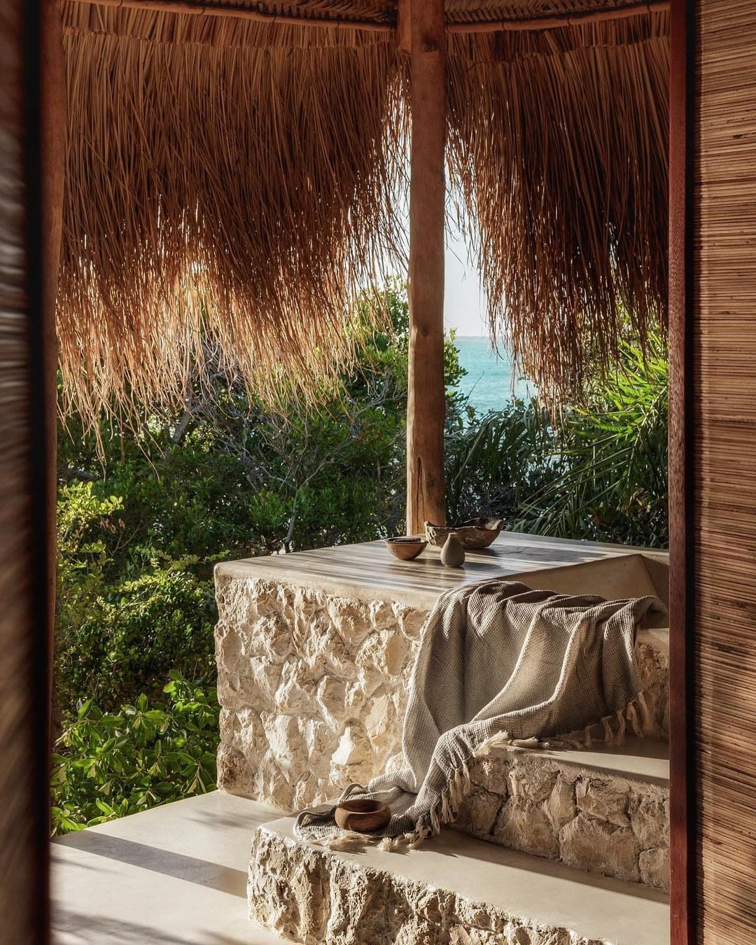 Sussurro, Africa — Coastal pavilion with thatched roof frame, stone bench, linen textile, bronze vessels, ocean view through vegetation.