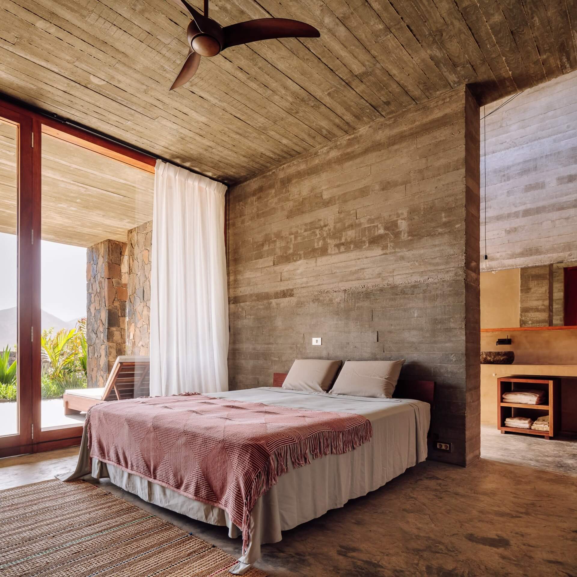 Barefoot Luxury, Africa — Concrete bedroom with timber ceiling fan, stone accent wall, and sliding glass doors opening to landscape view.