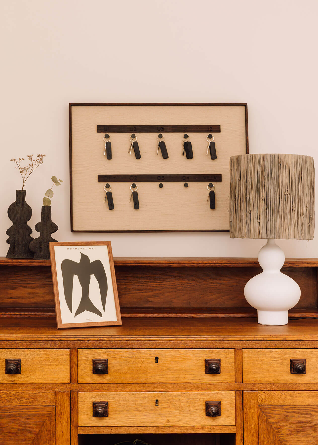 White Hotel, Europe — Mid-century wooden dresser styled with framed art, woven vessels, dried botanicals, and wall-mounted key organizer against neutral wall.