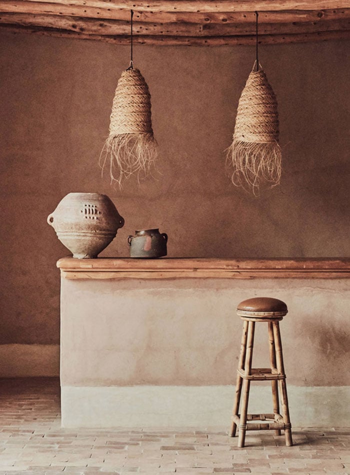 Berber Lodge, Africa — Rustic interior with woven pendant lights, terracotta walls, wooden counter, ceramic vessels, and wicker stool in minimalist earthen aesthetic.