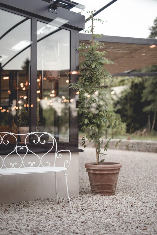 Le Barn, Europe — Illuminated pergola with white ornamental bench, climbing ivy in terracotta planter, gravel courtyard, warm ambient lighting at dusk.