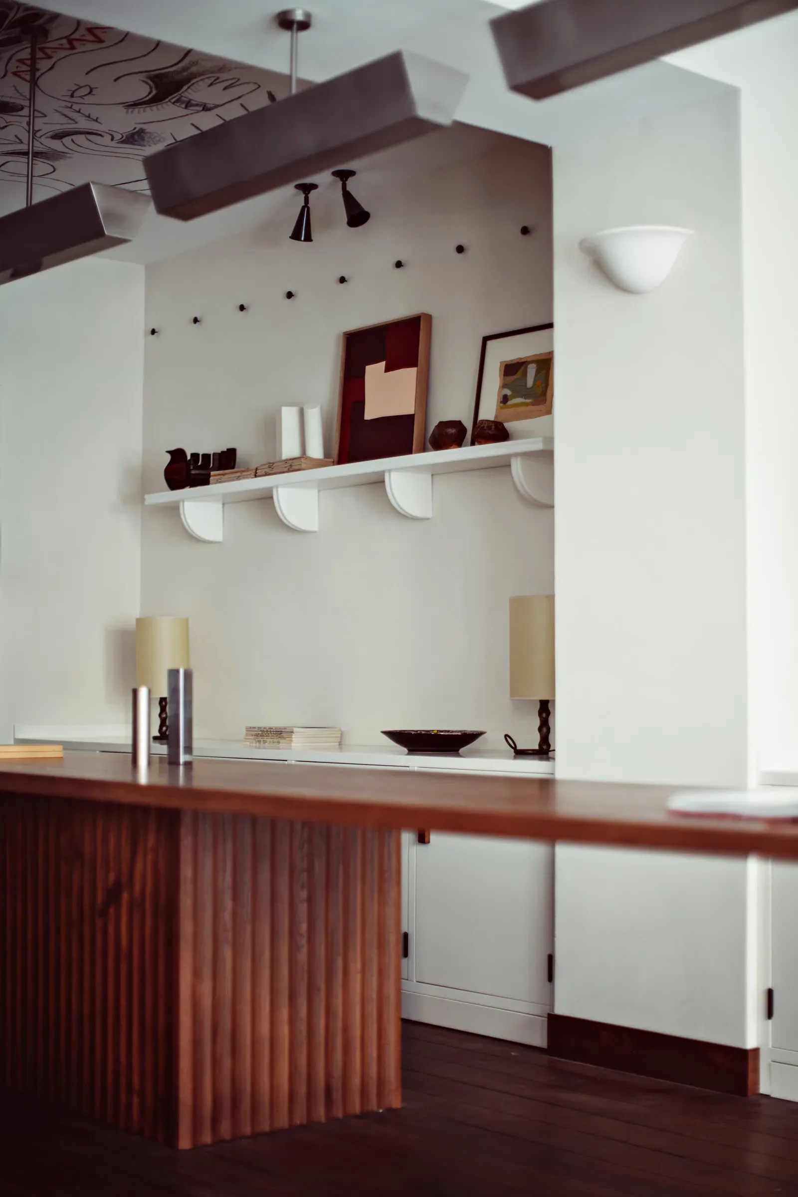 Académies et des Arts, Europe — Modern bar interior with dark wood counter, white wall-mounted shelving, exposed ceiling beams, burgundy accents, and contemporary pendant lighting.