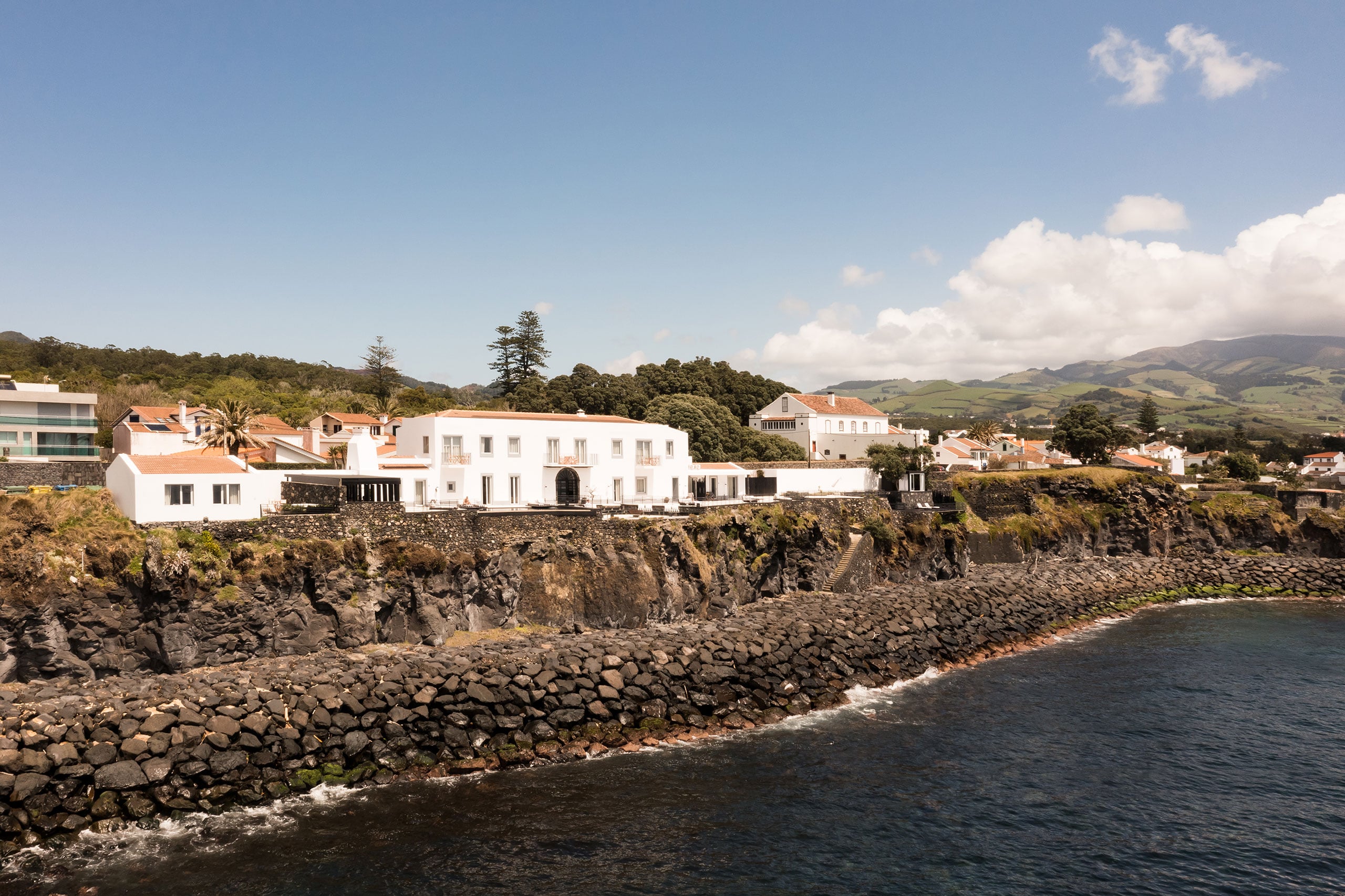 White Hotel, Europe — Whitewashed clifftop villa with terracotta roof overlooking basalt coastline, lush green hills beyond, contemporary luxury architecture on dramatic volcanic promontory.