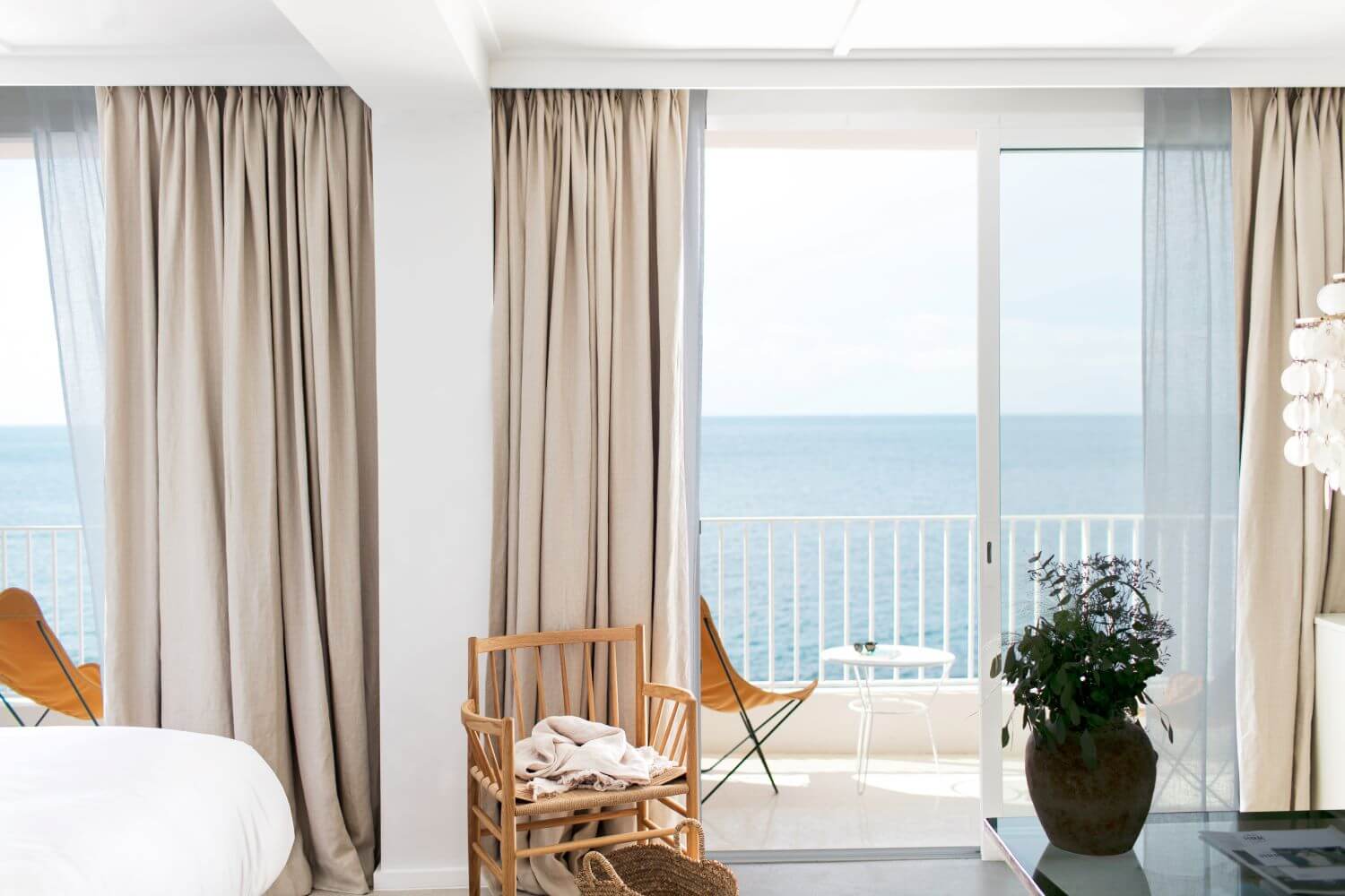 Les Roches Rouges, Europe — Coastal bedroom with floor-to-ceiling windows, beige linen curtains, wooden chairs, potted plant, and unobstructed sea views from balcony.