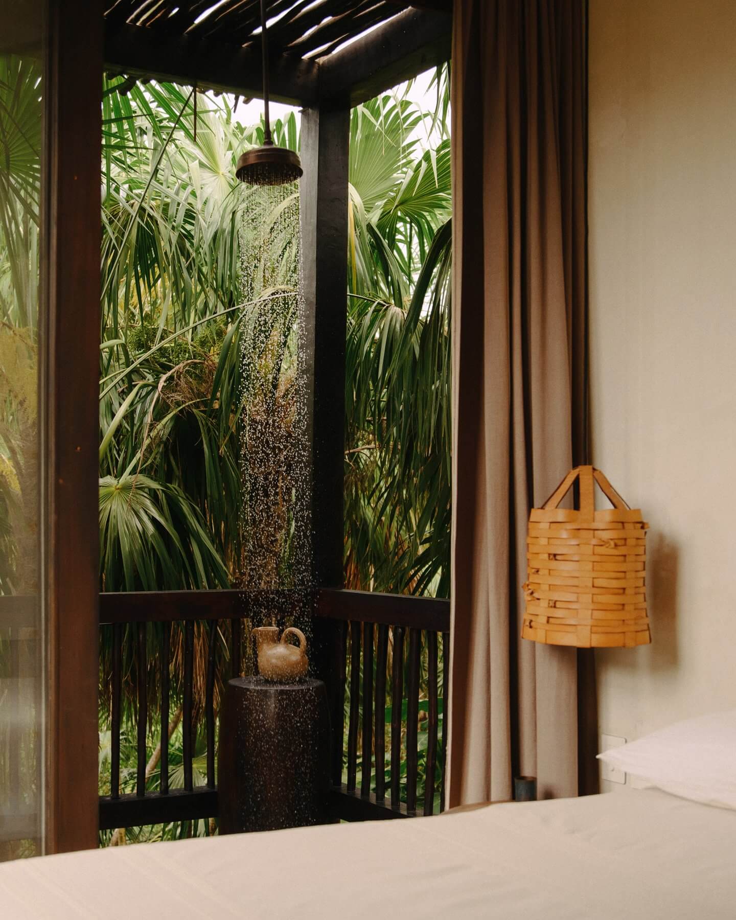 Tulum Treehouse, Mexico — Outdoor shower with rainfall showerhead surrounded by lush tropical palms and natural stone, framed by dark wooden doors and warm curtains.