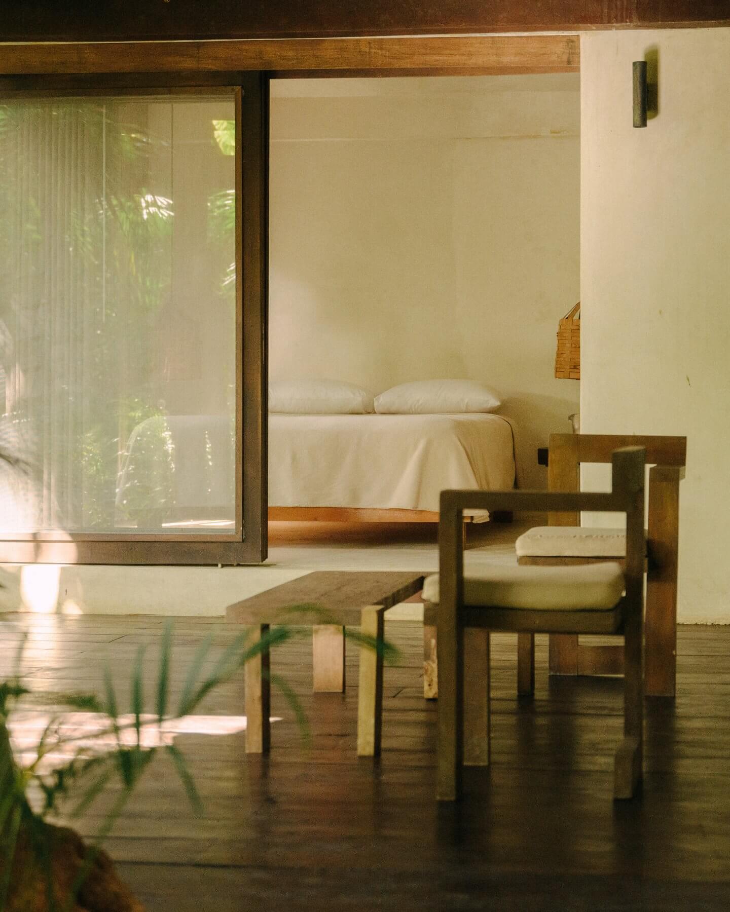 Tulum Treehouse, Mexico — Minimalist bedroom with wooden platform bed, cream linens, natural light through floor-to-ceiling glass doors, mid-century modern furniture, polished concrete floors.