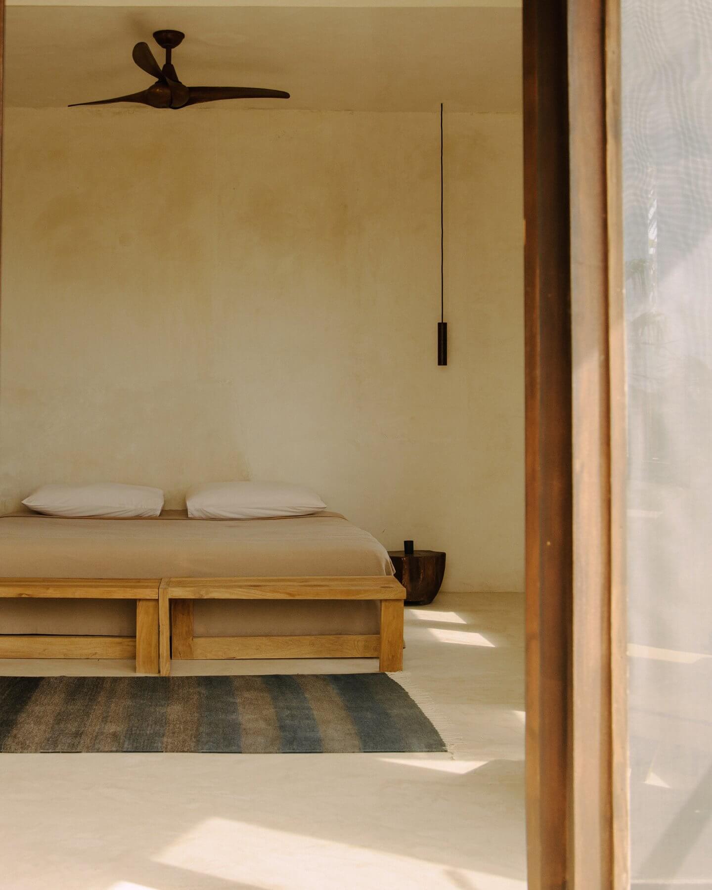 Tulum Treehouse, Mexico — Minimalist bedroom with wooden platform bed, white pillows, dark ceiling fan, pendant light, striped runner rug, and warm natural light through open wooden doorway.