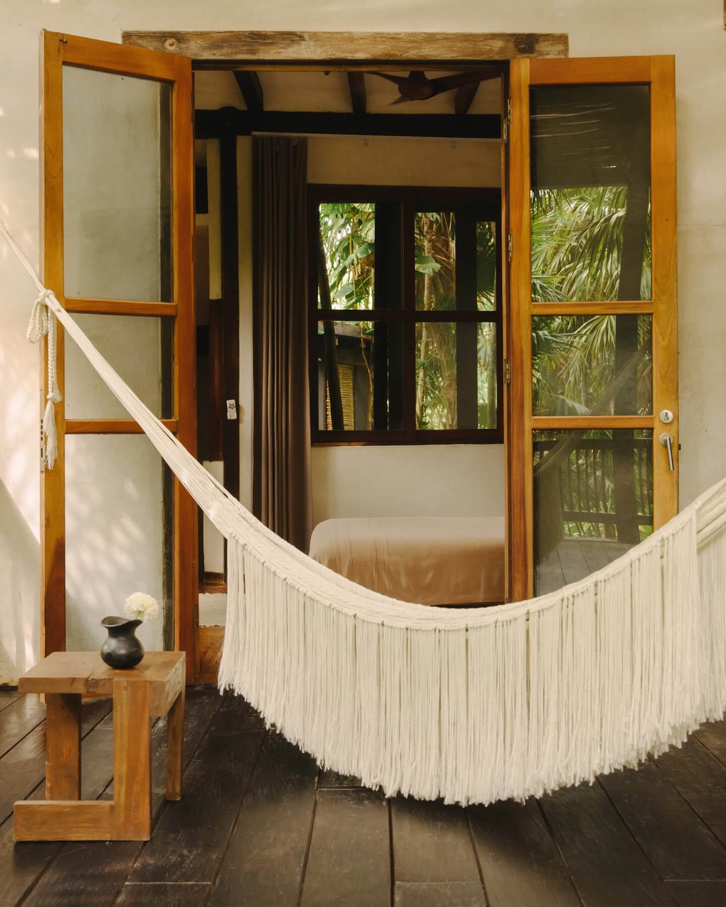 Tulum Treehouse, Mexico — Bedroom with wooden-framed glass doors opening to jungle canopy, white macramé hammock, dark wood flooring, and natural light filtering through palm foliage.
