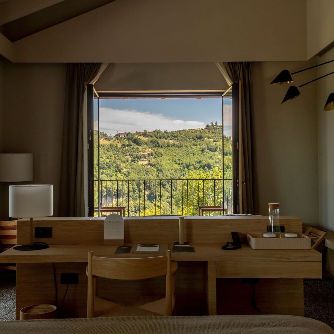Casa di Langa, Europe — Minimalist hotel room workspace with light-oak desks, panoramic doors opening to forested Tuscan hills, pendant lamps, neutral palette, natural daylight.