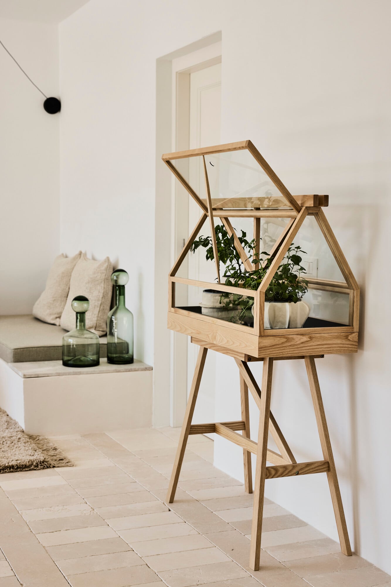 Villa Sessana, Italy — Wooden glass terrarium on tall legs with potted plants, minimalist interior with white walls, green glass bottles, and natural light.