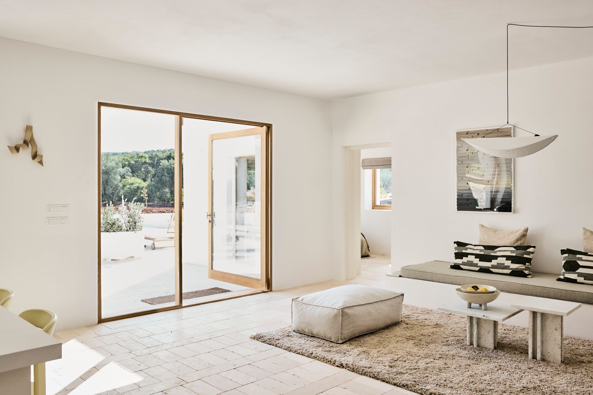 Villa Sessana, Italy — Minimalist living room with natural light, neutral palette, sliding glass doors opening to terrace, linen furnishings, and architectural simplicity.