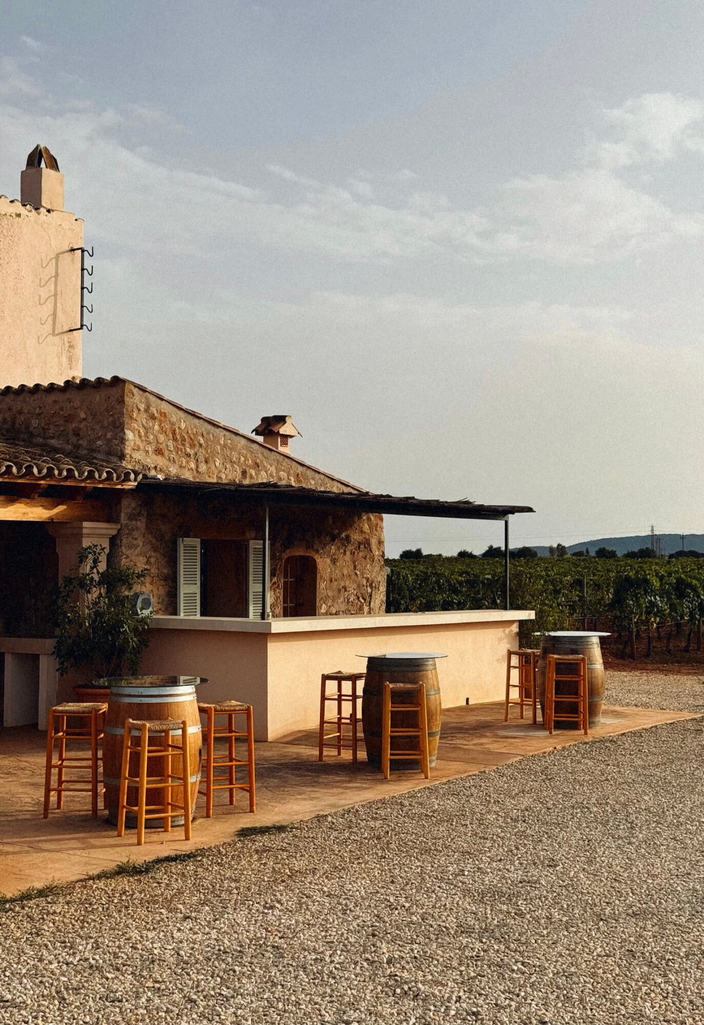 Sa Cabana, Balearic Islands, Spain — Rustic stone winery with terracotta courtyard, barrel seating, and vineyard views under Mediterranean light.